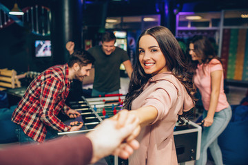 Beautiful yong woman look on caerma and smile. She stand in playing room. Model hold man's hand. Her friends stand at table soccer and play.