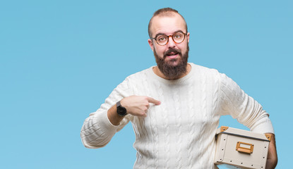 Young hipster man wearing glasses, moving holding moving box over isolated background with surprise face pointing finger to himself