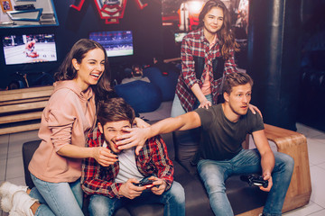 Two guys playing game on sofa in room. One of them hold hand in front of opponent's face. Young women laughing. They stand behind men.