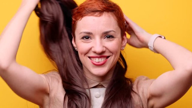 Funny changing style and emotions. Beautiful woman in classic wig with long brown hair positively taking off it. Girl turns stylish redhead pixie haircut. Female casual fashion on yellow background.