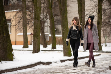 Two young stylish woman walks in snowy winter park. Positive emotions on their faces
