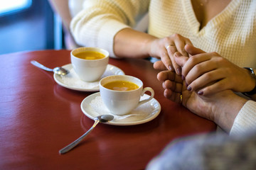 man and woman hold hands, close up of couple's arms on red wooden table in restaurant with two white cups with tea on it. date in cafe