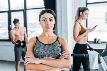 attractive athletic african american girl standing with crossed arms and smiling at camera in gym