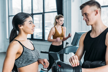 sportive young man and woman talking and looking at each other in gym, sportswoman training on treadmill behind