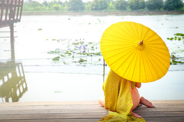 Beautiful woman wearing a Thai dress holding a yellow umbrella Sit back at the balcony of the waterfront.