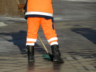 Street cleaning, municipal worker in boots and orange uniform sweep the street with a broom. Concept of unskilled labor, preparing for spring in city