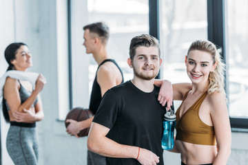 Fototapeta na wymiar happy young man and woman in sportswear standing together and smiling at camera in gym