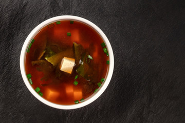 A photo of a bowl of miso shiru soup with tofu, scallions, and wakame seaweed, shot from the top on a black background with copy space