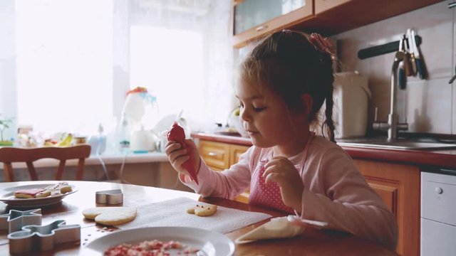Little girl preparing shortbread cookies in the kitchen. He enthusiastically draws icing. Festive atmosphere. 4K video