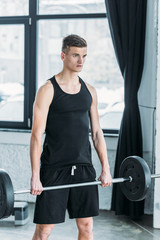 concentrated young athlete lifting barbell and looking away in gym