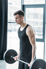 concentrated young sportsman lifting barbell and looking away in gym