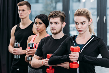 multiethnic sporty young friends holding dumbbells and looking away in gym