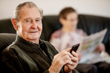 Senior sitzt zuhause im Wohnzimmer und surft auf seinem smart phone im Internet