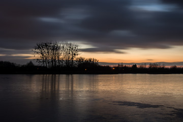 Fototapeta na wymiar Cloudy dark clouds after sunset on a frozen lake and bouncing trees in the ice