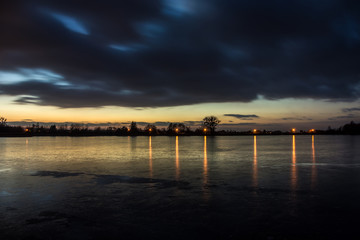 Evening clouds after sunset on a frozen lake and reflecting lights in the ice