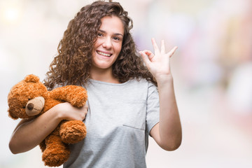 Young brunette girl holding teddy bear over isolated background doing ok sign with fingers, excellent symbol