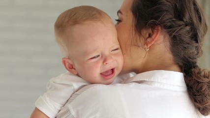 Young mother soothes a crying little baby at home in the bedroom. Close-up of a little newborn baby crying, mom calms him down.