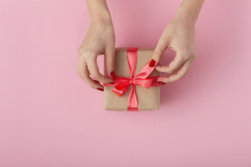 girl unties a ribbon bow on a gift box with hands, female with present wrapped in decorative paper on pink background, top view, concept holiday