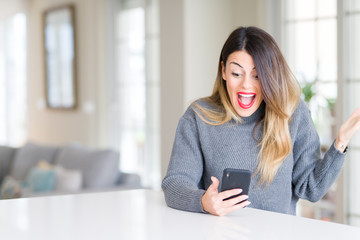 Young beautiful woman using smartphone at home very happy and excited, winner expression celebrating victory screaming with big smile and raised hands