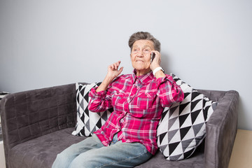 Theme old person uses technology. Mature happy joy smile active gray hair Caucasian wrinkles woman sitting home in living room on the sofa and using a mobile phone, calling and talking on the phone