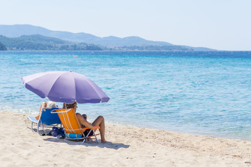 Man and woman sitting on small fold-out chairs on the beach under purple sunshade with a view of the blue sea water