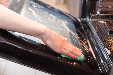 Housework and housekeeping concept. Scrubbing the stove and oven. Close up of female hand with green sponge cleaning the glass door of kitchen oven.