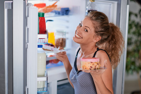 Woman taking cakes while standing in front of opened fridge.