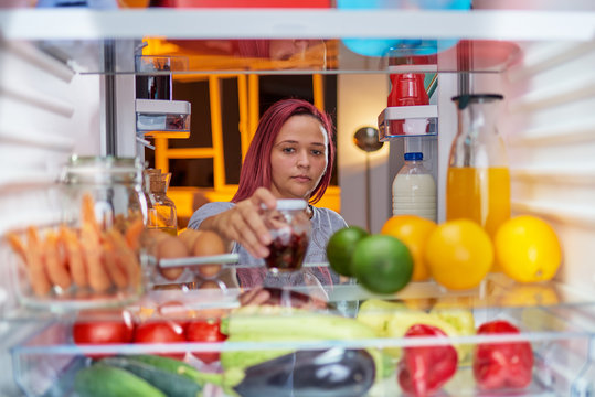 Woman standing in front of opened fridge  late at night and looking something to eat. Fridge full of groceries. Picture taken from the iside of fridge.