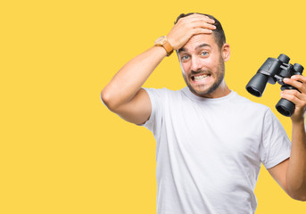 Young handsome man looking through binoculars over isolated background stressed with hand on head, shocked with shame and surprise face, angry and frustrated. Fear and upset for mistake.