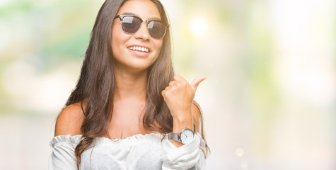 Young beautiful arab woman wearing sunglasses over isolated background smiling with happy face looking and pointing to the side with thumb up.