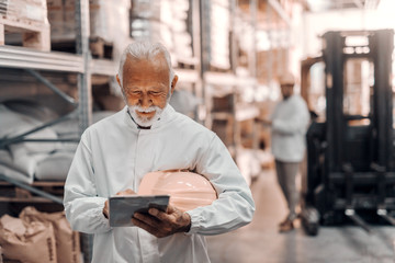 Happy dedicated young Caucasian worker with helmet under the armpit using tablet while standing in warehouse. In background worker and forklift.