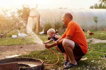 Grandfather playing with his grandson while crouching and spraying with hose. In background ducks and hens. Countryside exterior.