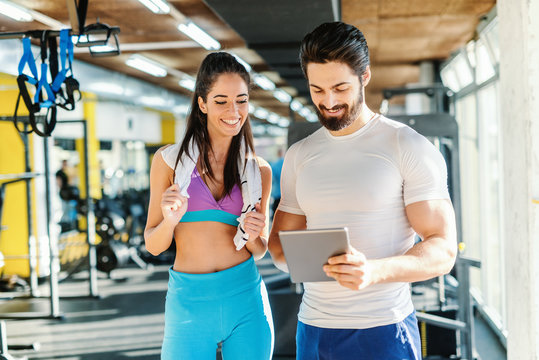 Bearded smiling personal trainer showing woman results of training on tablet. Woman standing next to him and looking at tablet. Gym interior.