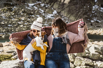 .Two young women, covered with a blanket, on a sunny winter morning enjoying a day on the mountain with their cute little dog. Lifestyle.