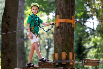 Young child boy in safety harness and helmet attached with carbine to cable on rope way in park.