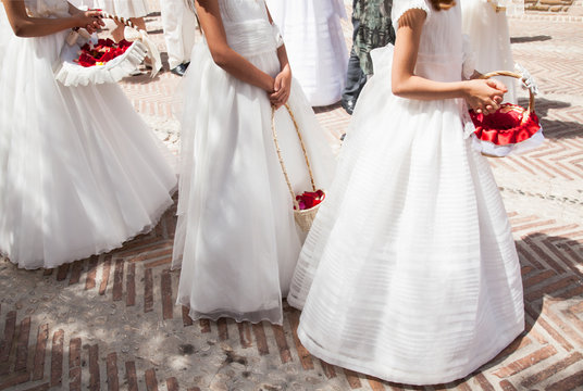 niñas llevando vestidos de comunión  blancos y largos con cesto con flores rojas
