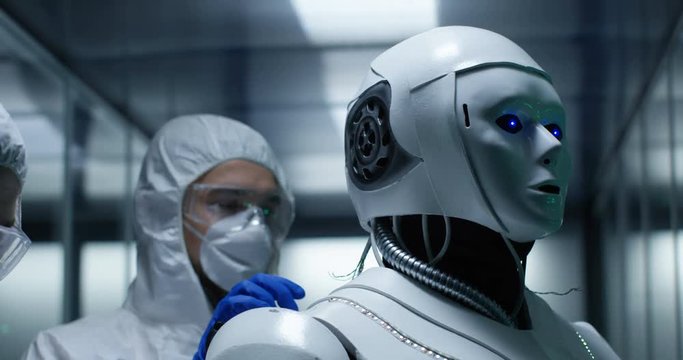 Medium shot of an engineer in protective suits try to turn on the robot fixing wires on robot control while colleague is monitoring in laboratory