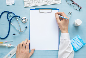 The top view of the doctor’s hands is writing down a prescription or medical report on a blank sheet of paper. On the table are stethoscope and other attributes. Copy space.   - obrazy, fototapety, plakaty