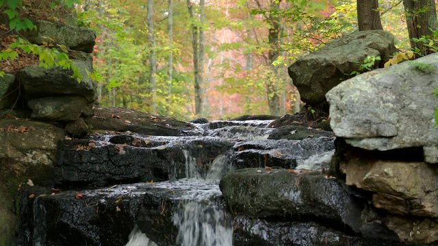 Spectacular peaceful shot of a small waterfall as the water cascades over dark mossy rocks in a foggy misty forest in during autumn with colorful fall leaves. Slow motion. 4K.