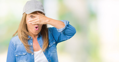 Beautiful middle age woman wearing sport cap over isolated background peeking in shock covering face and eyes with hand, looking through fingers with embarrassed expression.