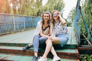 Two hipster girls in cool glasses are sitting on the old wooden bridge. Young fashionable sisters are having fun outdoors. Summer photoshoot for friends in the nature outdoors. Lifestyle concept.