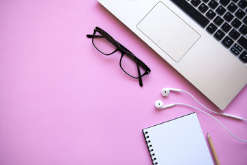 Workspace in a trendy pink color. Many different items including a laptop, glasses, notebook, and headphones are on the table. There is a place for text.