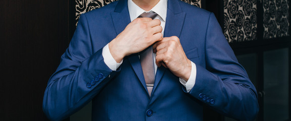 Groom is wearing the blue wedding suit. Stylish man is holding his tie in hands. Celebration day...