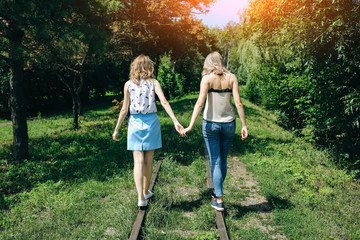 Naklejka na ściany i meble Two hipster girls are walking on the railway in the green park. Same sex couple is holding hands together. Travel style life concept. Support and friendship relationships.