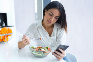 Pretty young woman using her mobile phone while eating salad in the kitchen at home.