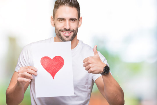 Handsome young man holding card with red heart over isolated background happy with big smile doing ok sign, thumb up with fingers, excellent sign