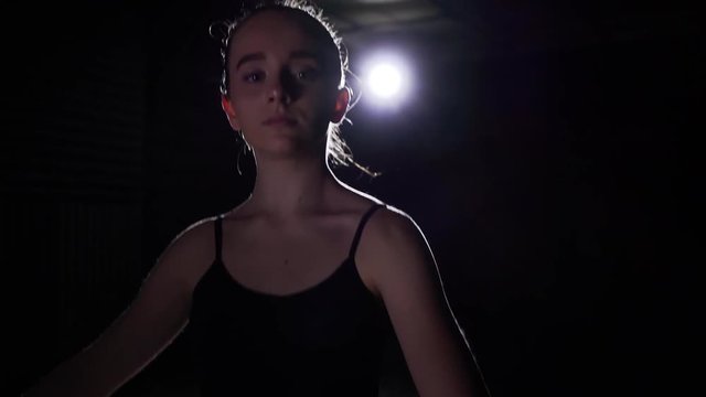 Young ballet dancer standing in spotlight on black background in studio. Ballerina shows classic ballet pas. Slow motion.