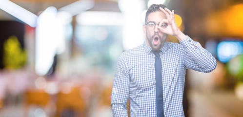 Young business man wearing glasses over isolated background doing ok gesture shocked with surprised face, eye looking through fingers. Unbelieving expression.