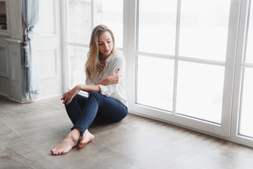 Fototapeta na wymiar Model blonde white shirt blue pants jeans sitting on wooden parquet floor by window.