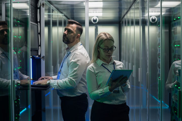 Diverse adult man and woman using tablet and laptop while diagnosing server hardware in modern data center
